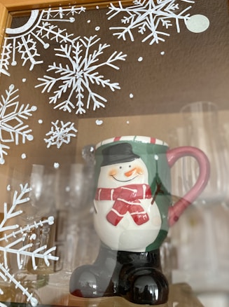 A ceramic mug designed in the shape of a cheerful snowman wearing a red scarf and a black hat. The mug has a pink handle, and it is set against a background featuring decorative white snowflake patterns. Glassware is visible in the background, providing a cozy winter or holiday atmosphere.