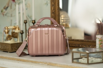A vintage-style room with a rose gold suitcase or handbag placed on a white tabletop. The background features an ornate gold-framed mirror, an antique rotary phone with golden accents, and decorative elements such as a candle holder and floral wallpaper. A glass box filled with pearls and a floral ceramic container are also present, adding to the elegant, retro vibe of the setting.