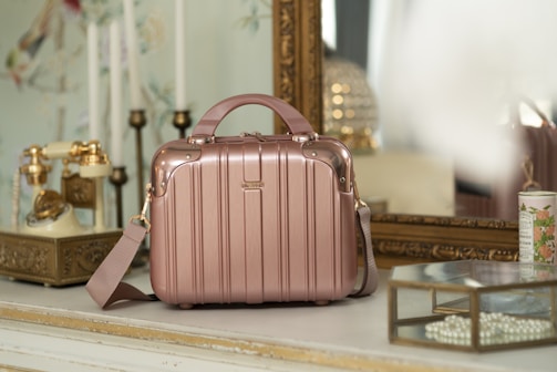 A vintage-style room with a rose gold suitcase or handbag placed on a white tabletop. The background features an ornate gold-framed mirror, an antique rotary phone with golden accents, and decorative elements such as a candle holder and floral wallpaper. A glass box filled with pearls and a floral ceramic container are also present, adding to the elegant, retro vibe of the setting.
