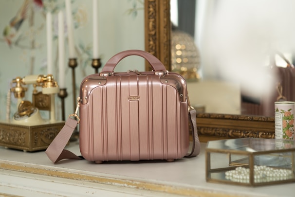 A vintage-style room with a rose gold suitcase or handbag placed on a white tabletop. The background features an ornate gold-framed mirror, an antique rotary phone with golden accents, and decorative elements such as a candle holder and floral wallpaper. A glass box filled with pearls and a floral ceramic container are also present, adding to the elegant, retro vibe of the setting.