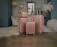 Four rose gold luggage bags of varying sizes are neatly arranged on a polished floor in a stylishly decorated room. The background features elegant hanging lamps, a white upholstered chair, a plush rug, ornate wallpaper, and a framed window with a view resembling the Eiffel Tower. A decorative feather display and a lace umbrella add to the sophisticated ambiance.