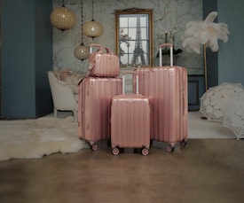 Four rose gold luggage bags of varying sizes are neatly arranged on a polished floor in a stylishly decorated room. The background features elegant hanging lamps, a white upholstered chair, a plush rug, ornate wallpaper, and a framed window with a view resembling the Eiffel Tower. A decorative feather display and a lace umbrella add to the sophisticated ambiance.