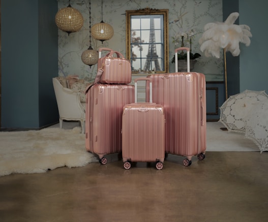 Four rose gold luggage bags of varying sizes are neatly arranged on a polished floor in a stylishly decorated room. The background features elegant hanging lamps, a white upholstered chair, a plush rug, ornate wallpaper, and a framed window with a view resembling the Eiffel Tower. A decorative feather display and a lace umbrella add to the sophisticated ambiance.