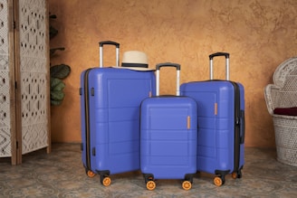 Three blue suitcases of varying sizes are arranged against a textured orange wall. The suitcases have black handles and orange wheels. A straw hat rests on the largest suitcase. Woven and wicker decorative elements are seen in the background, suggesting a travel or holiday theme.