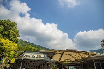 The image features a modern architectural structure labeled 'Penang Hill' surrounded by lush green trees under a clear blue sky with large, fluffy clouds. The foreground shows signage for pedestrian directions, indicating a bustling tourist area.