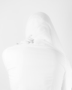 A minimalist image featuring a person wrapped in translucent white fabric, with their back partially turned and a hand gently placed on their shoulder. The composition creates a sense of softness and ethereality.