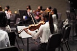 A group of musicians sitting in chairs, each holding a violin. The focus is on one musician in the foreground, wearing a white shirt, playing the violin and reading sheet music. Other musicians are visible in the background, also wearing formal attire and playing violins.