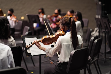 A group of musicians sitting in chairs, each holding a violin. The focus is on one musician in the foreground, wearing a white shirt, playing the violin and reading sheet music. Other musicians are visible in the background, also wearing formal attire and playing violins.