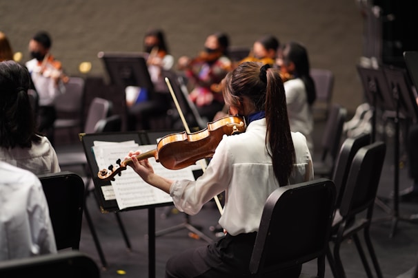 A group of musicians sitting in chairs, each holding a violin. The focus is on one musician in the foreground, wearing a white shirt, playing the violin and reading sheet music. Other musicians are visible in the background, also wearing formal attire and playing violins.