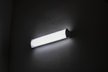 A fluorescent wall light fixture is mounted on a textured wall, emitting bright white light. The surrounding area is dimly lit, highlighting the fixture's sleek design.