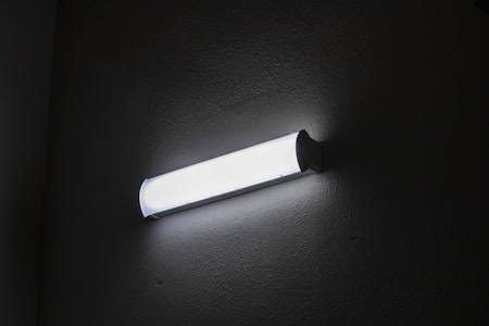 A fluorescent wall light fixture is mounted on a textured wall, emitting bright white light. The surrounding area is dimly lit, highlighting the fixture's sleek design.