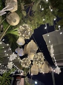 A whimsical outdoor setting featuring hanging decorative lanterns and fairy lights. The lanterns are varied in size and shape, including wicker and metallic designs, creating a warm, inviting atmosphere. Snowflake decorations add a festive touch amidst the green foliage, against a dark night sky.