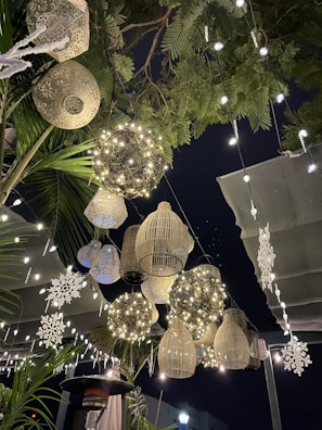 A whimsical outdoor setting featuring hanging decorative lanterns and fairy lights. The lanterns are varied in size and shape, including wicker and metallic designs, creating a warm, inviting atmosphere. Snowflake decorations add a festive touch amidst the green foliage, against a dark night sky.