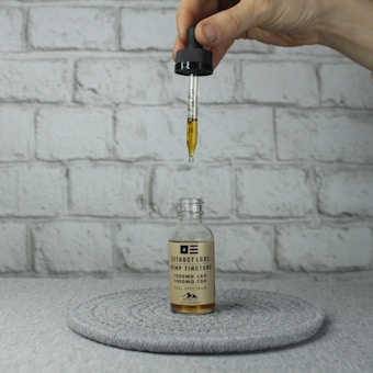 A hand holds a dropper above a glass bottle labeled 'Extract Labs Hemp Tincture', with a focus on the liquid being dispensed into the bottle. The setup is against a gray brick background, with the bottle placed on a round gray mat.