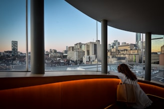 A person with long hair leans on a railing inside a curved, modern building with tall windows. The view outside reveals a city skyline during twilight, with various skyscrapers and an empty horizon in the distance. The interior of the building is well-lit with a warm glow, contrasting with the cooler colors of the sky and buildings outside.