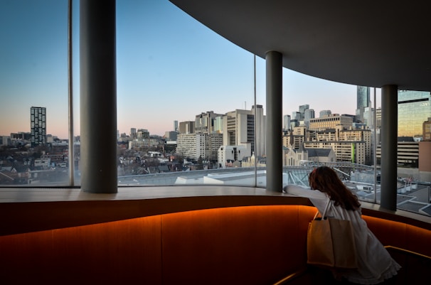 A person with long hair leans on a railing inside a curved, modern building with tall windows. The view outside reveals a city skyline during twilight, with various skyscrapers and an empty horizon in the distance. The interior of the building is well-lit with a warm glow, contrasting with the cooler colors of the sky and buildings outside.