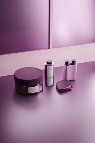 A collection of purple-toned cosmetic containers is arranged on a similarly colored surface. The set includes a round jar, a tall cylindrical bottle, a square container, and a rectangular holder with two bottles inside. The background features a gradient of purple shades.
