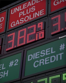 A large gas station price sign displays fuel prices with LED lights. The sign includes sections for unleaded gasoline, diesel, and other fuel types, with each section prominently showing prices in red and green LED numbers.