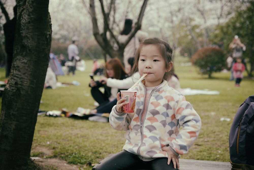 a little girl sitting on a park bench drinking a drink