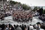 A group of performers wearing traditional attire stand in a circle, raising their arms towards the sky, in an outdoor setting. They are surrounded by a large audience sitting on steps, observing the performance. The scene is set in a natural environment with greenery and an ocean view in the background.