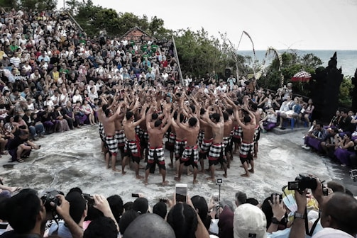 A group of performers wearing traditional attire stand in a circle, raising their arms towards the sky, in an outdoor setting. They are surrounded by a large audience sitting on steps, observing the performance. The scene is set in a natural environment with greenery and an ocean view in the background.