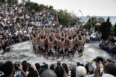 A group of performers wearing traditional attire stand in a circle, raising their arms towards the sky, in an outdoor setting. They are surrounded by a large audience sitting on steps, observing the performance. The scene is set in a natural environment with greenery and an ocean view in the background.