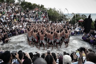 A group of performers wearing traditional attire stand in a circle, raising their arms towards the sky, in an outdoor setting. They are surrounded by a large audience sitting on steps, observing the performance. The scene is set in a natural environment with greenery and an ocean view in the background.