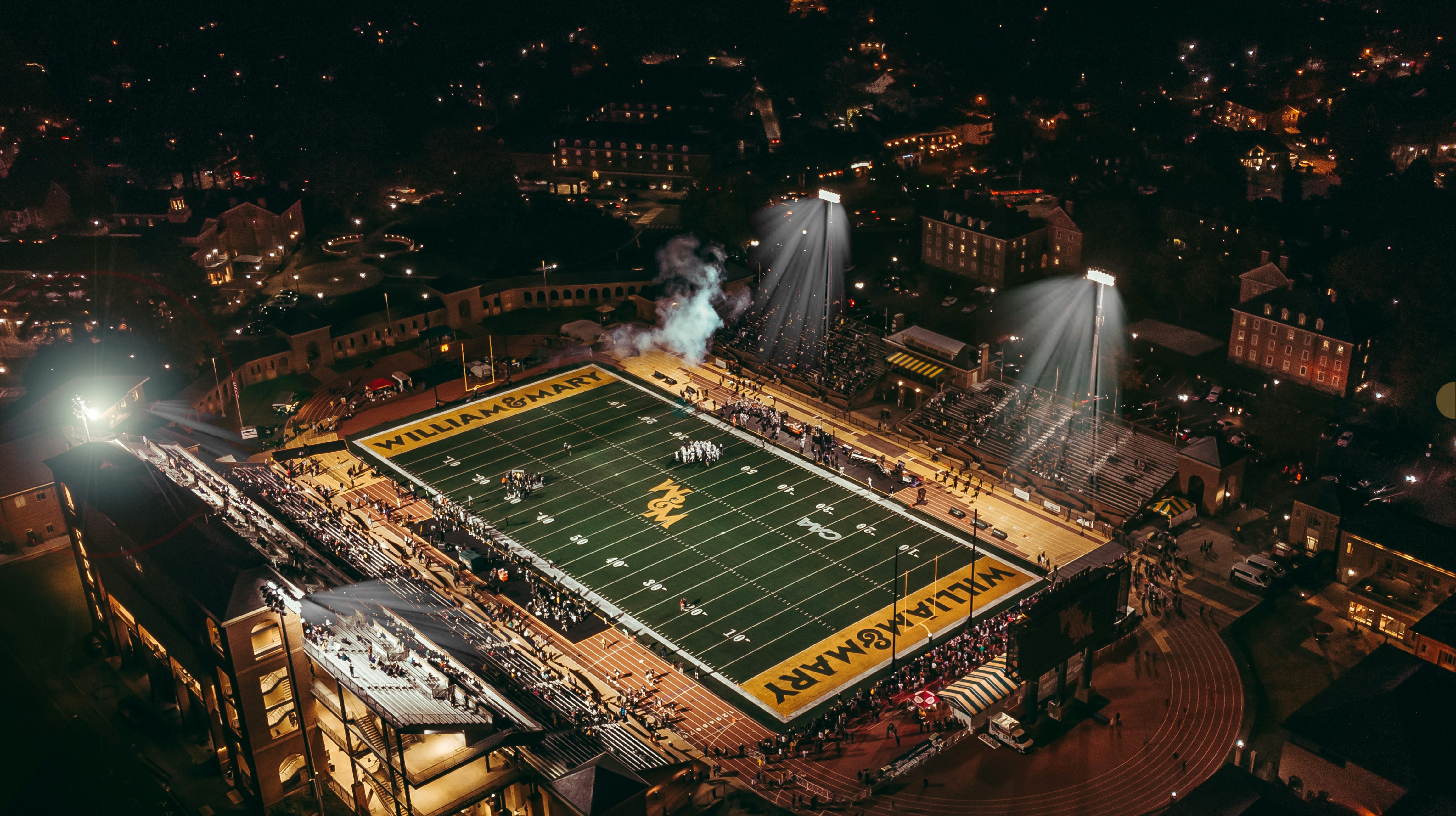 Stadium at night with dramatic floodlights, crowd atmosphere, sports field below, aerial cinematic perspective