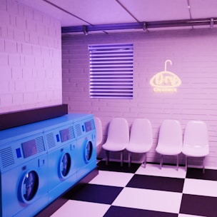 A brightly lit laundry room with a modern design. There are four blue washing machines lined up on the left side. On the right side, there are five white plastic chairs arranged in a row against a light pink brick wall. Above the chairs, a neon sign reading 'Dry Cleaners' is mounted on the wall. The floor has a black and white checkered tile pattern. Above the chairs, there's a window with horizontal blinds.