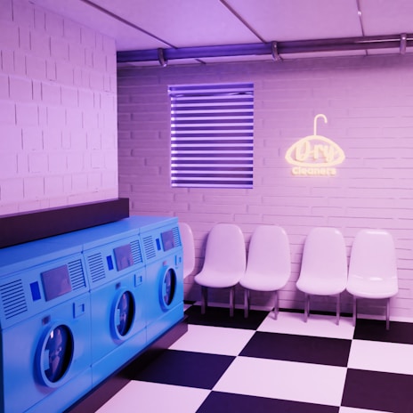 A brightly lit laundry room with a modern design. There are four blue washing machines lined up on the left side. On the right side, there are five white plastic chairs arranged in a row against a light pink brick wall. Above the chairs, a neon sign reading 'Dry Cleaners' is mounted on the wall. The floor has a black and white checkered tile pattern. Above the chairs, there's a window with horizontal blinds.
