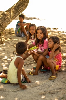 A group of children is sitting on a sandy beach, engaging in conversation under the shade of a large tree. They seem relaxed and are dressed in casual summer clothing.