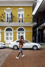 A person wearing a red shirt, denim shorts, and white sneakers walks along a brick sidewalk in front of a yellow two-story building featuring arched windows and a balcony with decorative iron railings. A sign on the upper floor indicates leasing information. A white car is parked on the street adjacent to the building. Another building with dark accents is partially visible on the right.