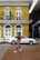 A person wearing a red shirt, denim shorts, and white sneakers walks along a brick sidewalk in front of a yellow two-story building featuring arched windows and a balcony with decorative iron railings. A sign on the upper floor indicates leasing information. A white car is parked on the street adjacent to the building. Another building with dark accents is partially visible on the right.