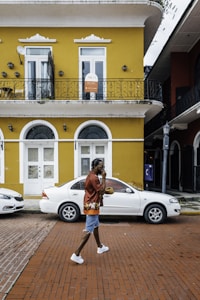 A person wearing a red shirt, denim shorts, and white sneakers walks along a brick sidewalk in front of a yellow two-story building featuring arched windows and a balcony with decorative iron railings. A sign on the upper floor indicates leasing information. A white car is parked on the street adjacent to the building. Another building with dark accents is partially visible on the right.