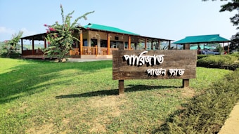 A wooden sign with white text written in a script is situated on a grassy area, surrounded by landscaped greenery. In the background, there is a building with a wooden porch and a green roof, accompanied by an open pavilion with a similar green roof. The sky is clear and blue, giving a view of a peaceful and idyllic setting.