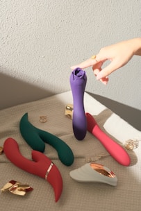 Various vibrantly colored personal massagers are arranged on a textured fabric surface. A hand with a ring is reaching towards a purple massager. The shadow and light suggest it is taken during the day.