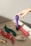 Various vibrantly colored personal massagers are arranged on a textured fabric surface. A hand with a ring is reaching towards a purple massager. The shadow and light suggest it is taken during the day.