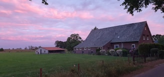 A rural landscape with a large brick house and an adjacent small barn set against a lush green field. The sky is painted with soft pink and purple hues, indicating a serene sunset or dawn. Trees and shrubs surround the buildings, adding to the tranquil, pastoral scene.
