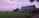 A rural landscape with a large brick house and an adjacent small barn set against a lush green field. The sky is painted with soft pink and purple hues, indicating a serene sunset or dawn. Trees and shrubs surround the buildings, adding to the tranquil, pastoral scene.