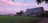 A rural landscape with a large brick house and an adjacent small barn set against a lush green field. The sky is painted with soft pink and purple hues, indicating a serene sunset or dawn. Trees and shrubs surround the buildings, adding to the tranquil, pastoral scene.