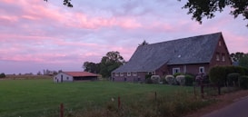 A rural landscape with a large brick house and an adjacent small barn set against a lush green field. The sky is painted with soft pink and purple hues, indicating a serene sunset or dawn. Trees and shrubs surround the buildings, adding to the tranquil, pastoral scene.