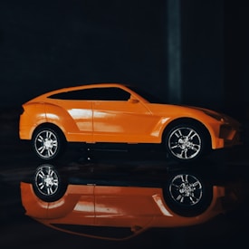 A toy model of a sleek orange sports car is positioned against a dark background. The car is highly detailed with visible reflections on a glossy surface below, enhancing its vibrant appearance.