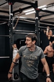 A person with a focused expression is in a gym environment, wearing a gray t-shirt with a graphic design. In the background, there are exercise equipment and other individuals engaged in workouts, including a person performing a pull-up.
