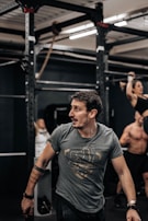A person with a focused expression is in a gym environment, wearing a gray t-shirt with a graphic design. In the background, there are exercise equipment and other individuals engaged in workouts, including a person performing a pull-up.