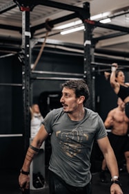 A person with a focused expression is in a gym environment, wearing a gray t-shirt with a graphic design. In the background, there are exercise equipment and other individuals engaged in workouts, including a person performing a pull-up.