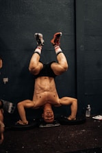 A person is performing a headstand against a dark wall, with their legs bent at the knees and feet resting on the wall. They are wearing athletic shoes, shorts, and colorful socks, with visible muscles on their upper body. Nearby, there is a water bottle and some papers on the floor.