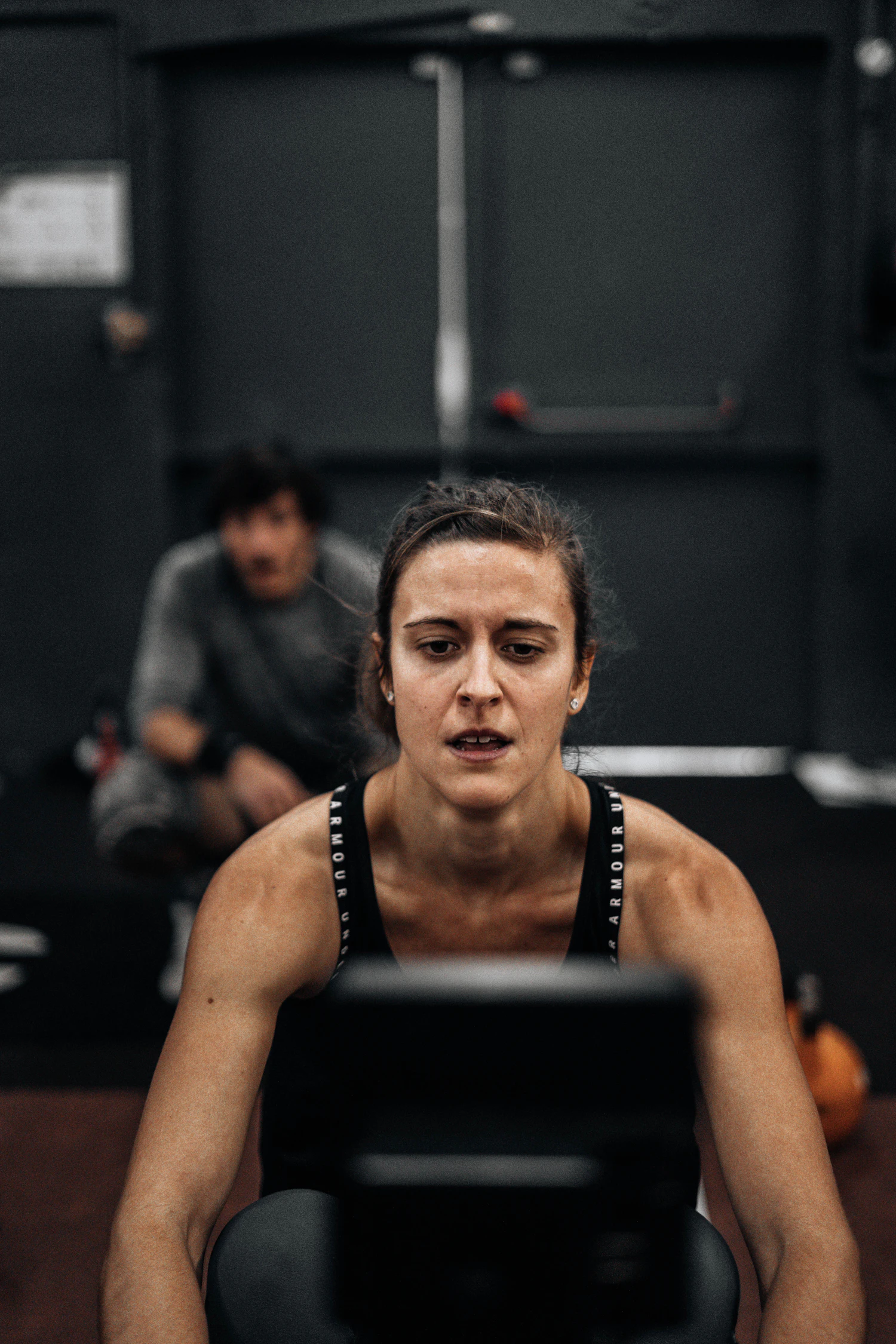 A woman pulling hard on a rowing handle in a dark room, jaw set, a second person behind her