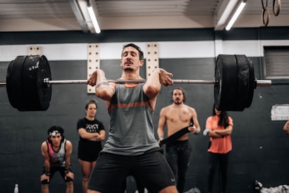 A man is performing a weightlifting exercise, holding a heavy barbell across his shoulders. He is in a gym environment with several other individuals in the background, some watching and others seemingly engaged in their own activities. The gym has a utilitarian atmosphere with dim lighting and visible structural elements.