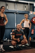 A group of people in an indoor gym setting, with two women standing and a man squatting and appearing to be shouting encouragement. They are dressed in workout clothes, surrounded by gym equipment such as weights and a red fire extinguisher in the background.