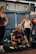 A group of people in an indoor gym setting, with two women standing and a man squatting and appearing to be shouting encouragement. They are dressed in workout clothes, surrounded by gym equipment such as weights and a red fire extinguisher in the background.